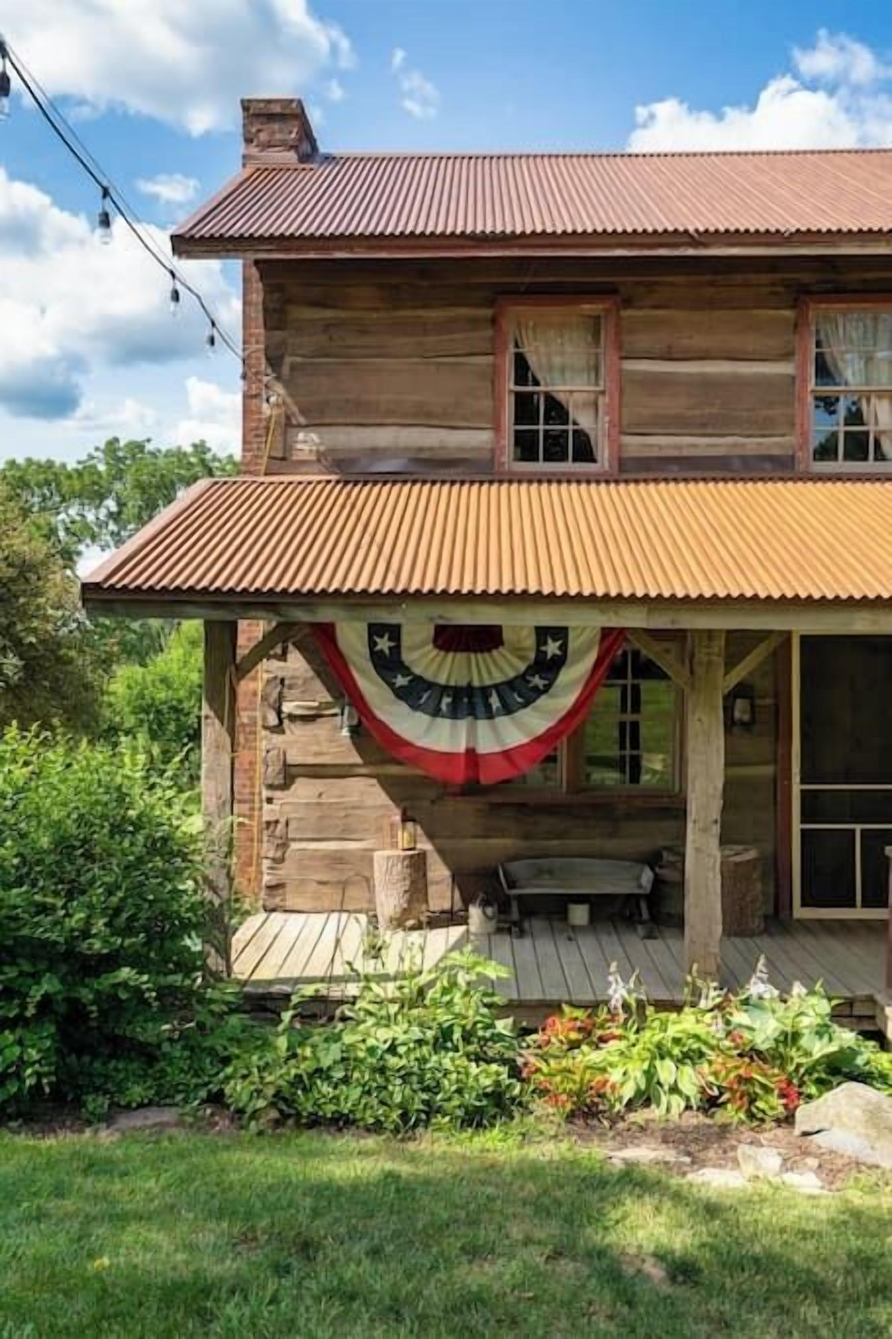 A two-story log cabin with a porch, a flag, and a garden.