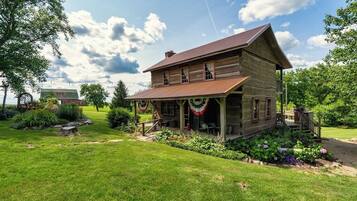 Un chalet rustique en rondins de bois, doté d’un porche, entouré d’un jardin bien entretenu et sous un ciel dégagé.