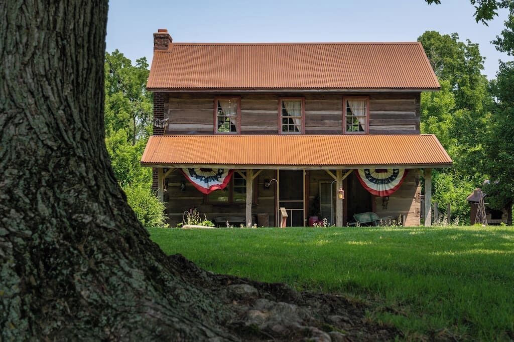 A two-story log cabin with a porch, surrounded by a grassy yard and trees.