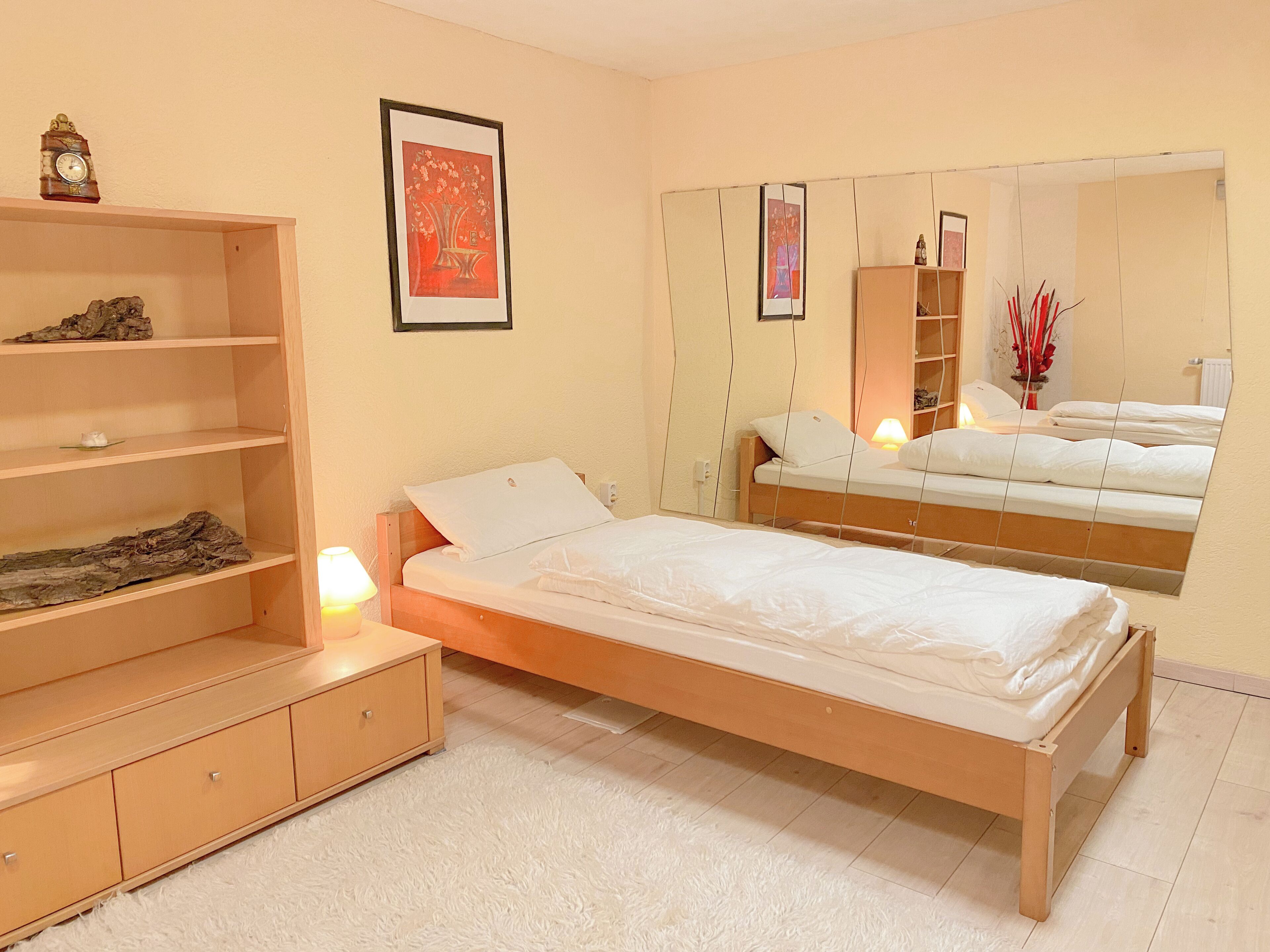 A bedroom with a single bed, a wooden dresser, a mirror, and a shelf with decorative items.