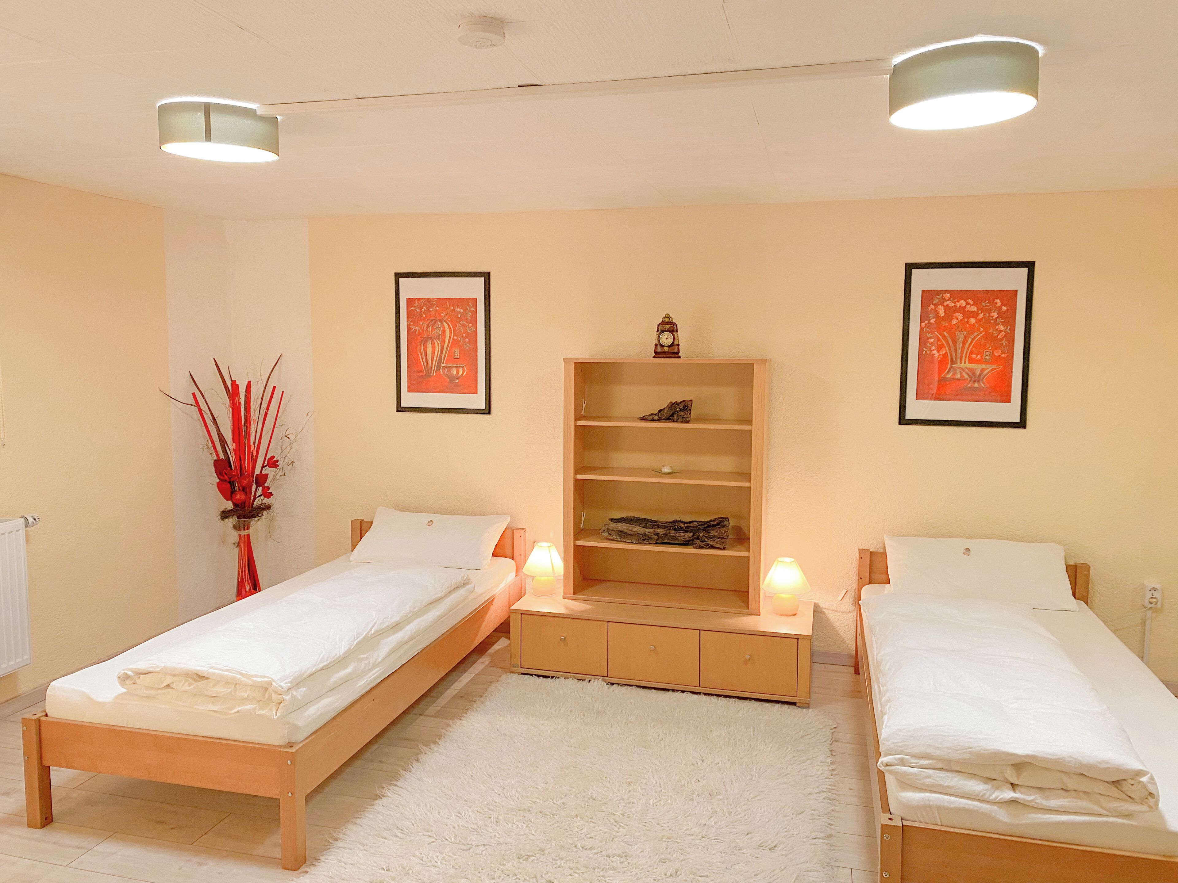 A hotel room with two single beds, a wooden nightstand with a lamp, a shelf with decorative items, and two framed pictures on the wall.