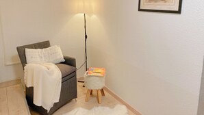 A cozy room with a grey armchair, a white throw blanket, a small stool with a magazine, a floor lamp, and a framed picture on the wall.
