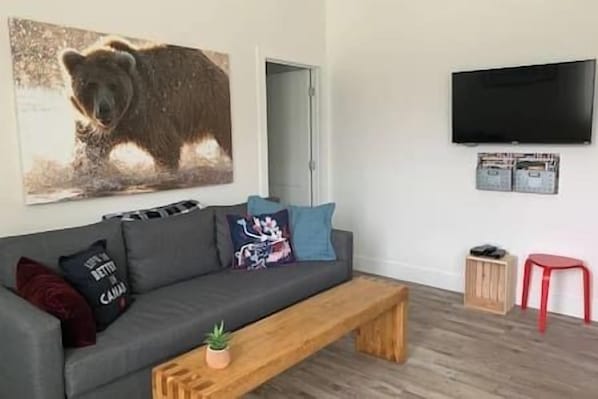 A living room with a grey sofa, a wooden coffee table, a bear painting, a television, and a small red stool.