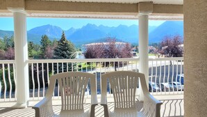 A covered outdoor area with two plastic chairs and a wooden table, overlooking a mountainous landscape with trees and buildings. - Mountain View Condo with Sofabed and Aircon (Fairmont Hot Springs)