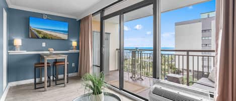 A modern living room with a large window, a flat-screen TV, a dining area, and a balcony with a view of the ocean.