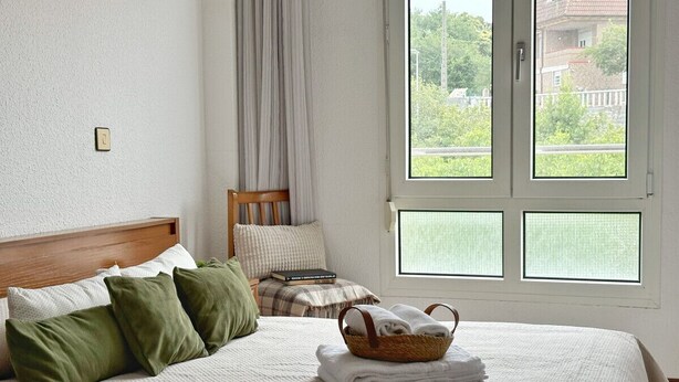 Un lit bien fait, avec une tête de lit, des oreillers et un panier rempli de serviettes pliées, et une vue sur la verdure à travers la fenêtre.