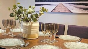 A dining table set with plates, glasses, and cutlery, accompanied by a vase of green foliage, in a room with a large scenic painting on the wall.