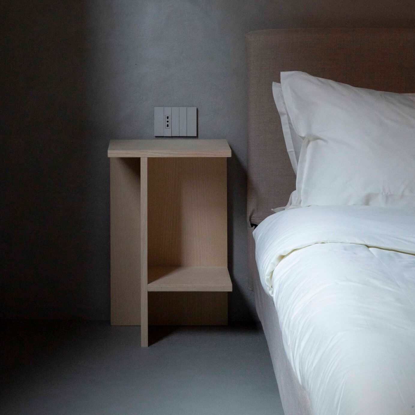A minimalist bedroom with a bed, a nightstand, and a wall-mounted light switch.