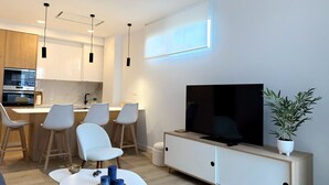 Un soggiorno moderno con una TV a schermo piatto, una zona pranzo con sedie e una cucina sullo sfondo.