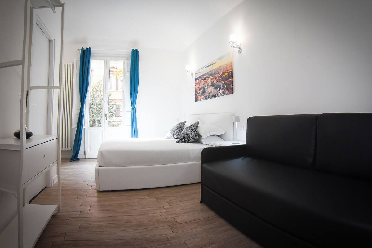 Un dormitorio con una cama, un sofá negro, un tocador blanco y un cuadro en la pared.