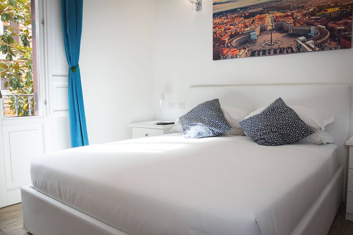 Una cama bien hecha con ropa de cama blanca y almohadillas estampadas, una mesita de noche blanca con un control remoto, y un cuadro de un paisaje urbano en la pared.