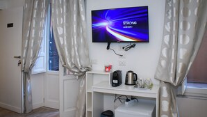 Habitación de hotel con una televisión montada en la pared mostrando un protector de pantalla, una cafetera, un hervidor y un mini refrigerador sobre un escritorio blanco.