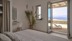 A bedroom with a bed, a chair, a potted plant, and a view of the sea through an open door.