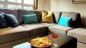 Un canapé modulable avec des coussins turquoise et jaunes, une petite table sur laquelle se trouve une assiette de fruits et un verre d’une boisson rouge, et une fenêtre laissant entrer la lumière naturelle.