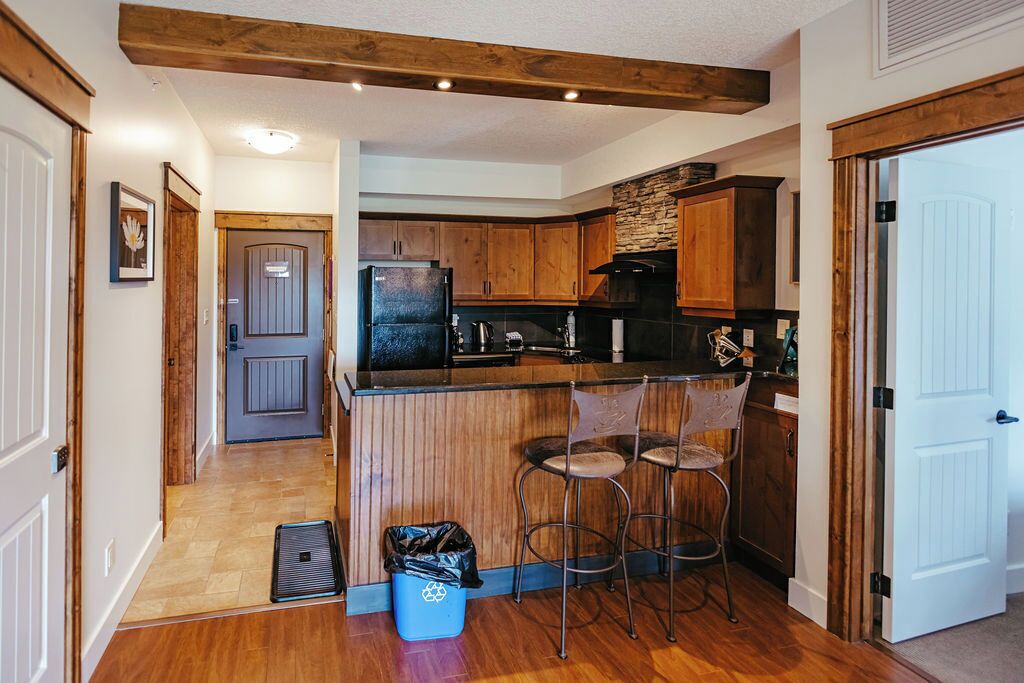 A kitchen with wooden cabinets, a black refrigerator, and a stone backsplash. There are two bar stools and a trash bin.