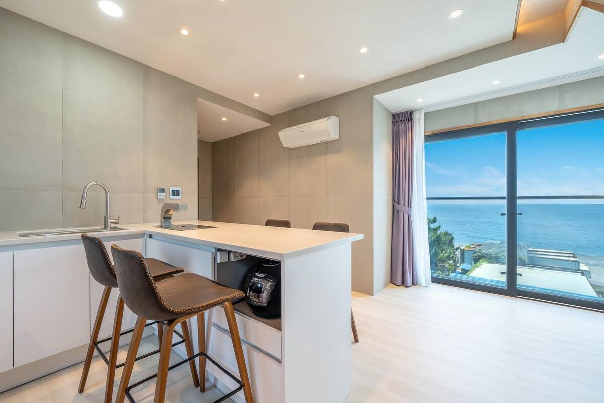 A modern kitchen with a central island, bar stools, and a view of the sea through large windows.