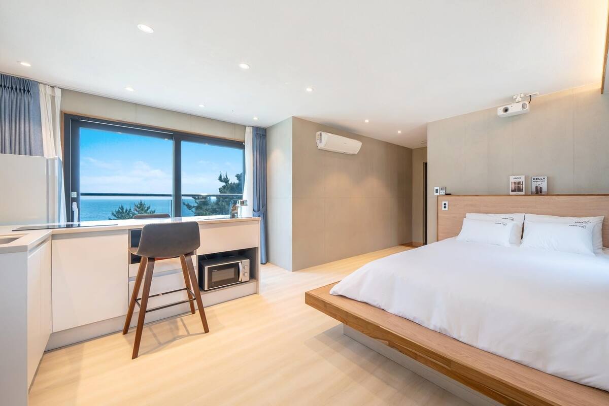 A modern hotel room with a large bed, a desk with a chair, a microwave, and a view of the sea.