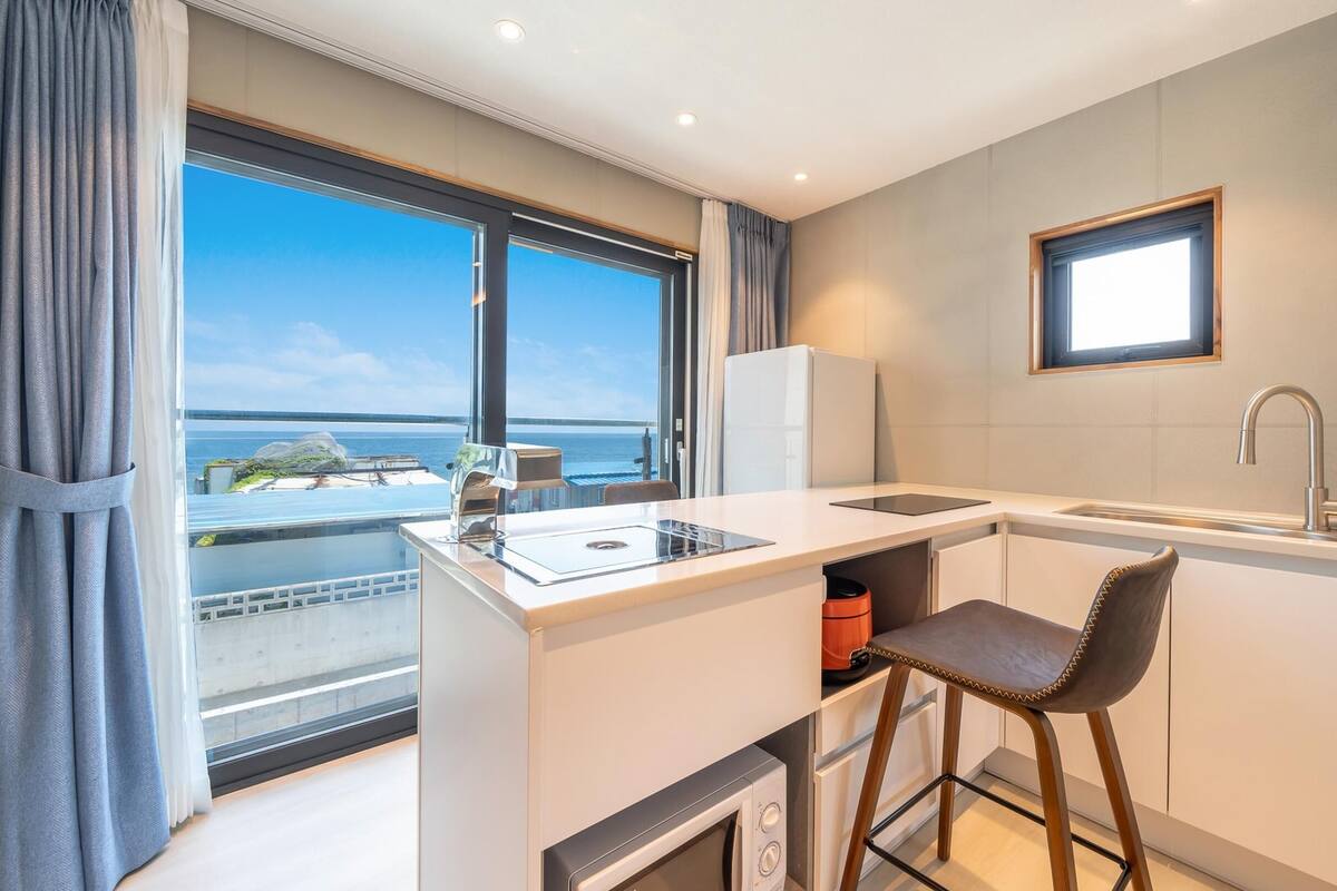 A modern kitchen with a view of the sea, a built-in oven, a sink, and a bar stool.