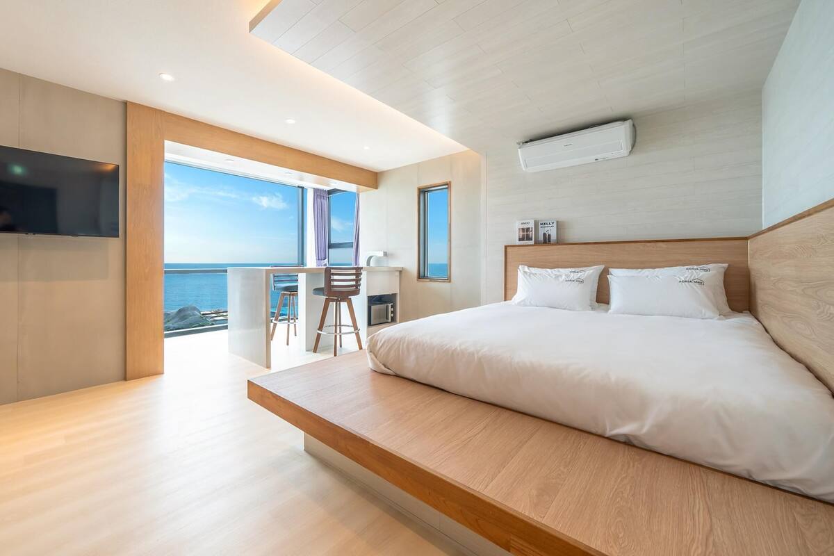 A modern bedroom with a large bed, a TV, and a balcony with a view of the sea.