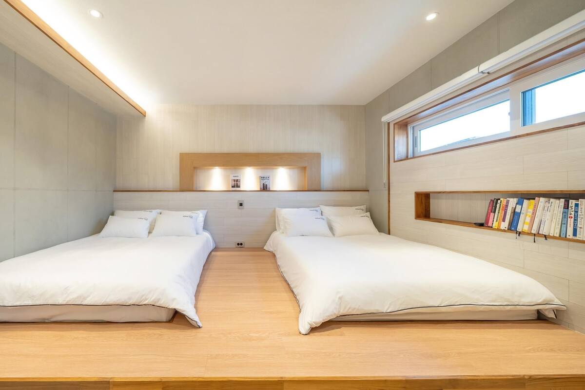 A modern hotel room with two single beds, a wooden floor, a built-in bookshelf, and a large window.