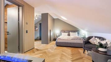 A modern bedroom with a herringbone wood floor, a bed with grey bedding, a grey armchair, and a bedside table with a lamp.