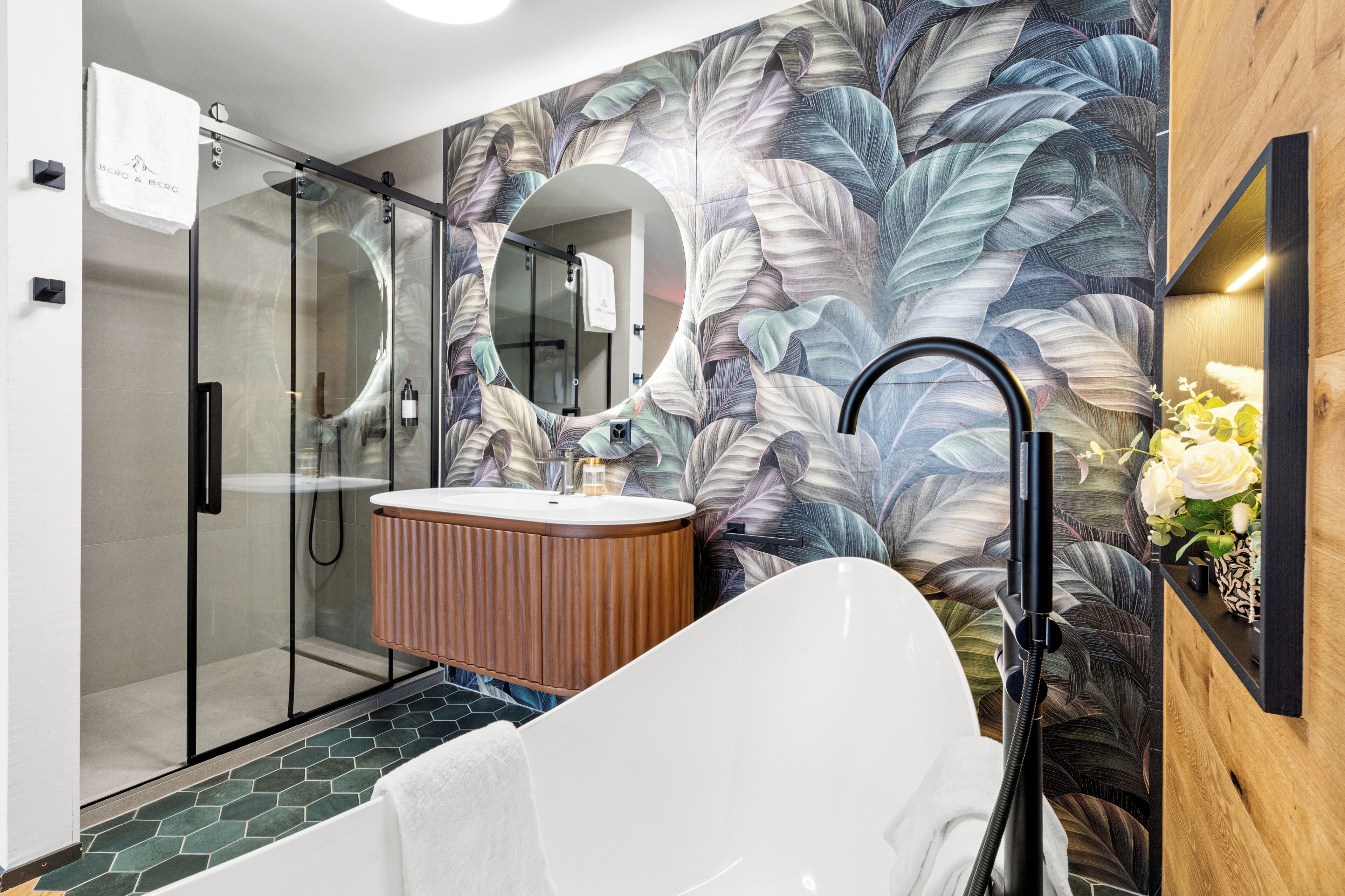 A modern bathroom with a glass shower enclosure, a freestanding bathtub, a wooden vanity with a round mirror, and a floral-patterned wall.
