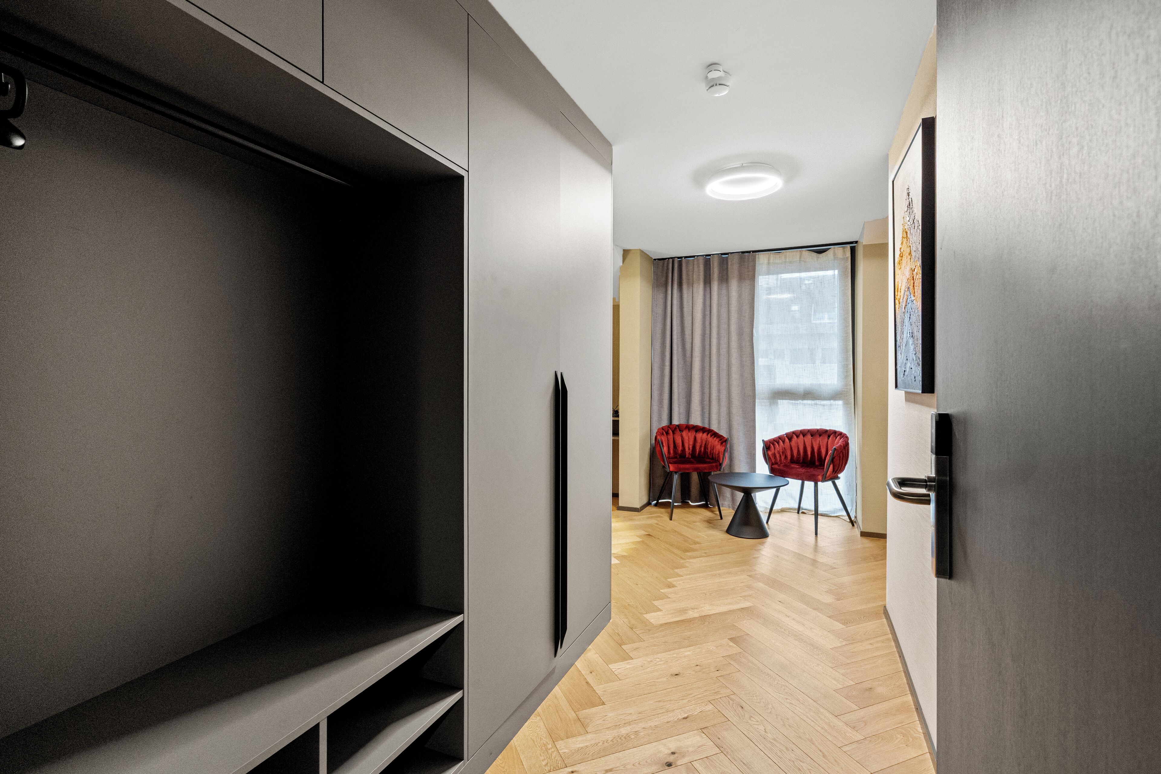 Una habitación de hotel moderna con un armario grande, una zona de comedor con dos sillas rojas y vistas a la ciudad a través de amplias ventanas.