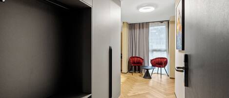 Una habitación de hotel moderna con un armario grande, una zona de comedor con dos sillas rojas y vistas a la ciudad a través de amplias ventanas.