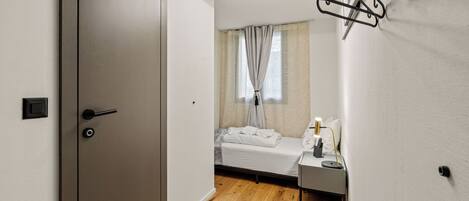 A bedroom with a wooden floor, a bed, a bedside table with a lamp, and a window with curtains.