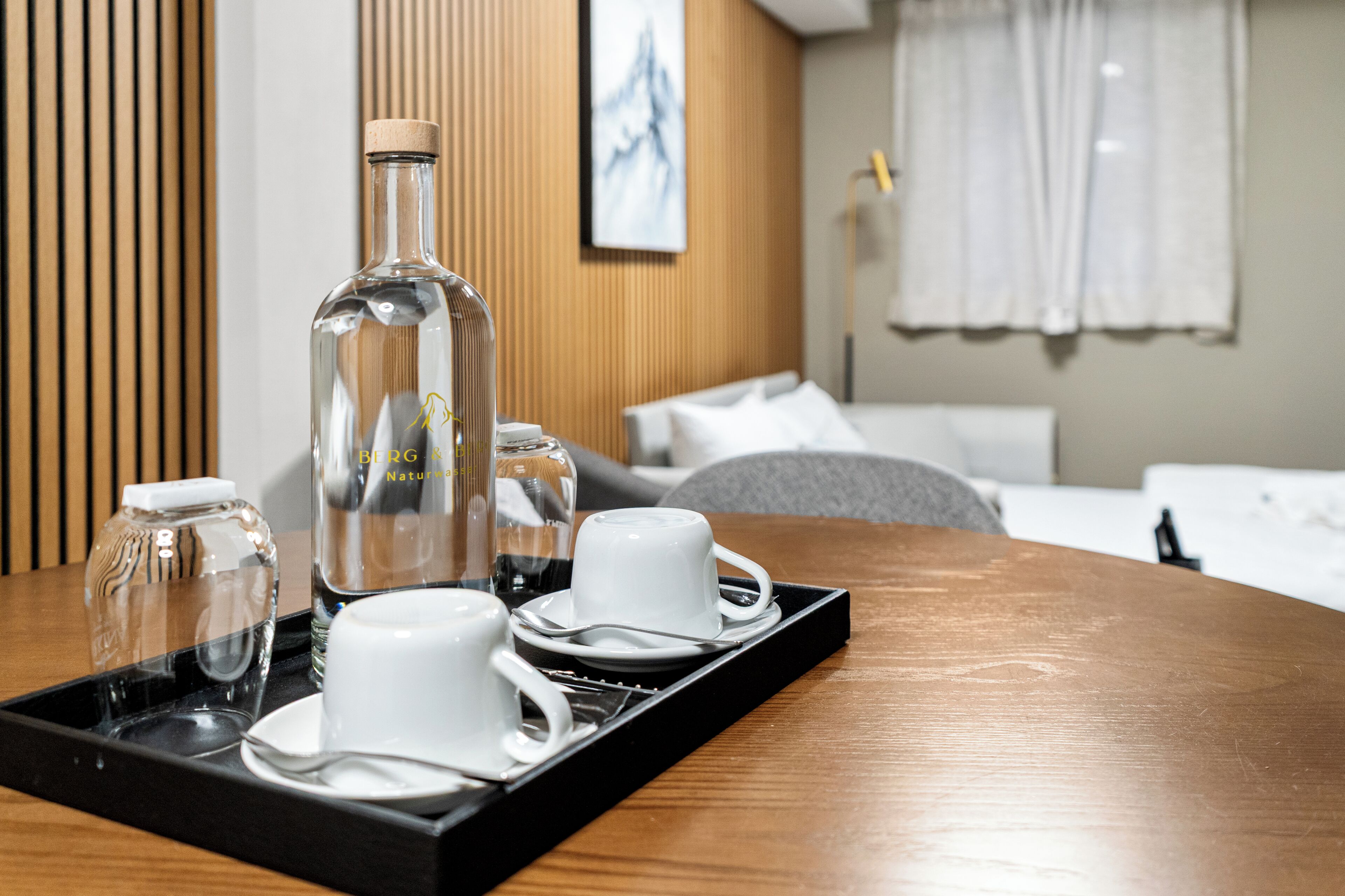 Habitación de hotel con panel de pared de madera, cama con ropa blanca, mesita de noche con una bandeja que contiene una botella, dos tazas y un vaso.