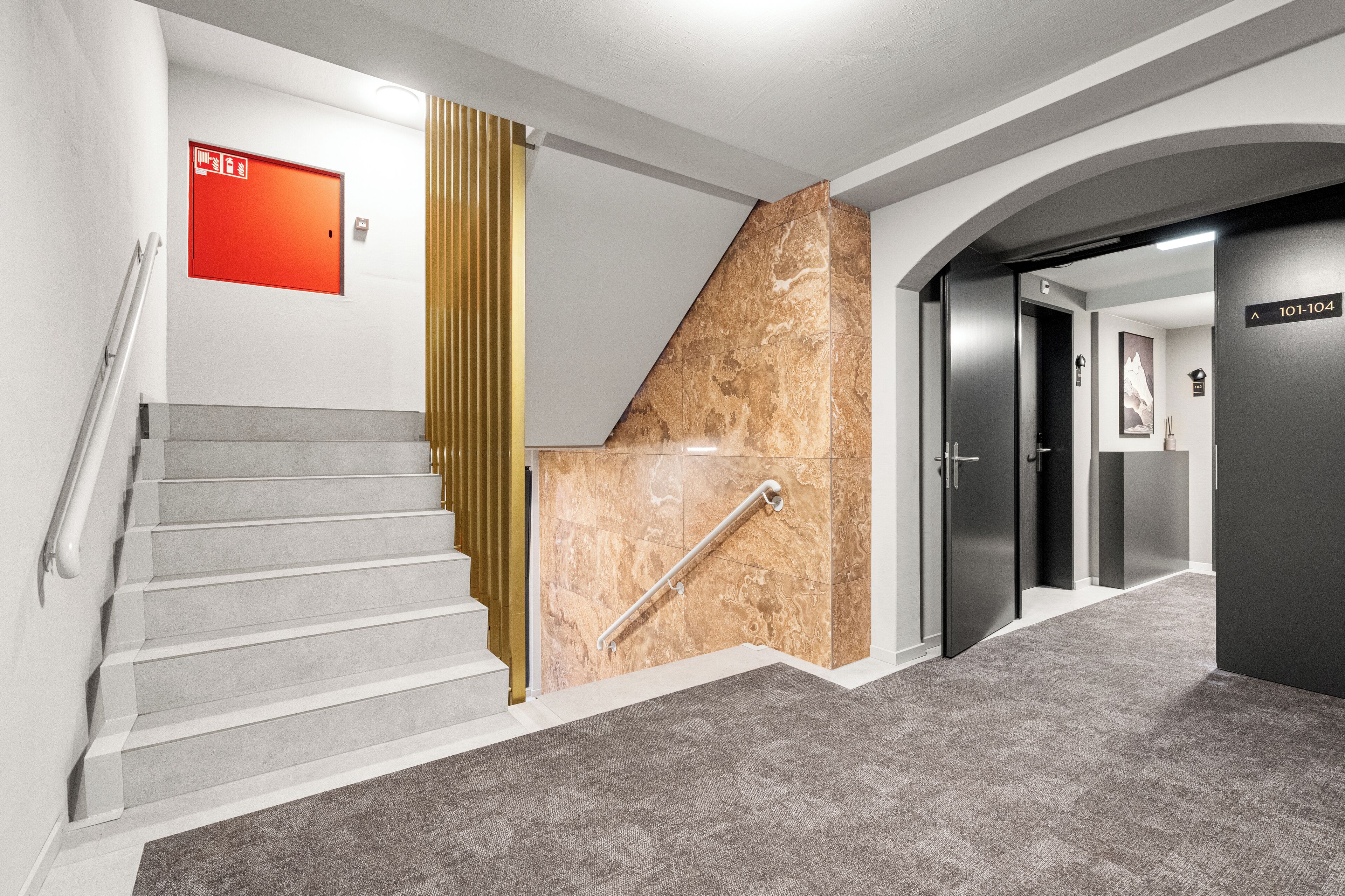 A modern hotel lobby with a marble staircase, a red emergency exit sign, and a hallway leading to elevators.