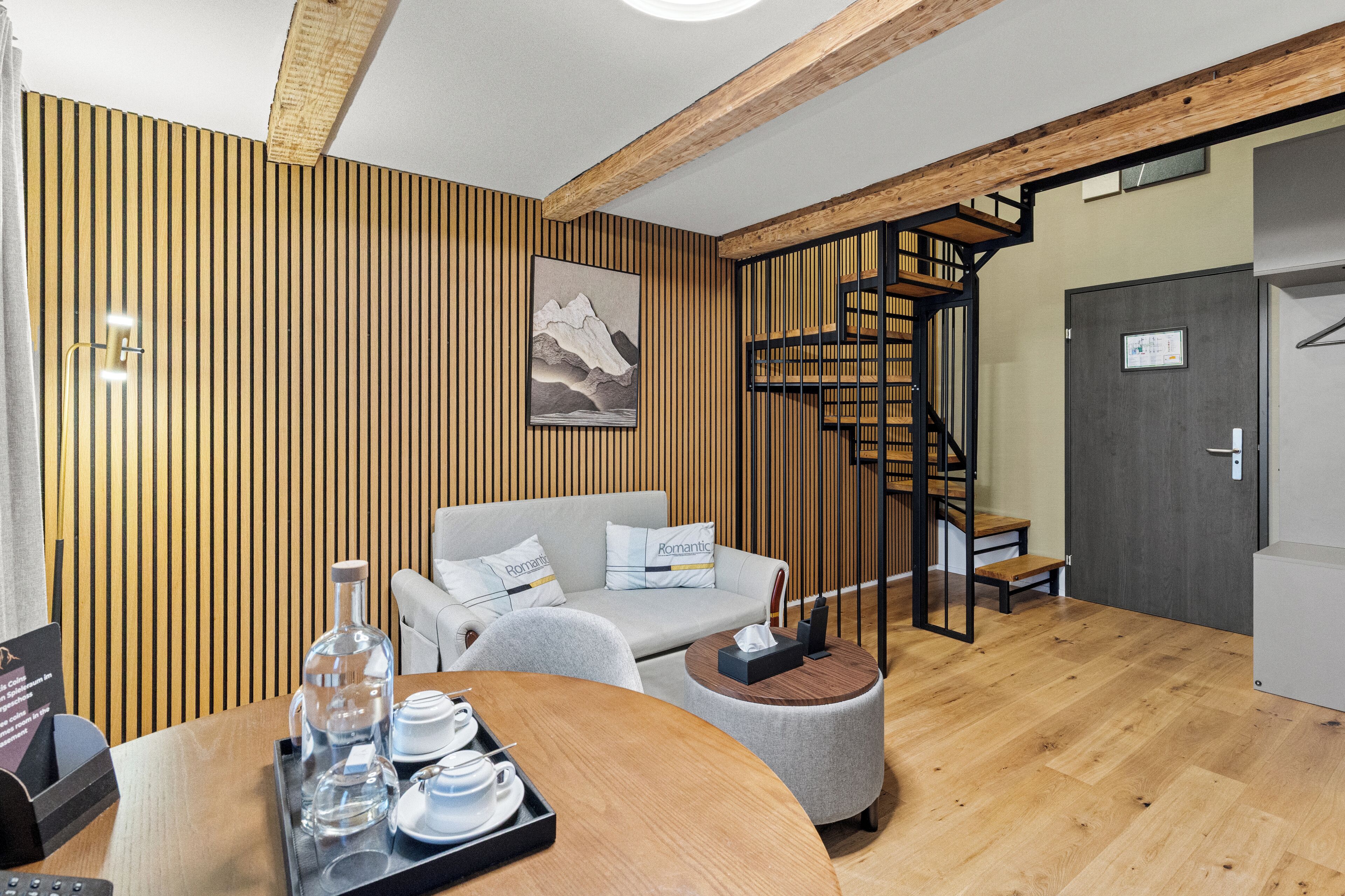 A modern hotel room with a wooden panel wall, a sofa, a round table with a tray of tea and glasses, and a staircase with a wooden railing.