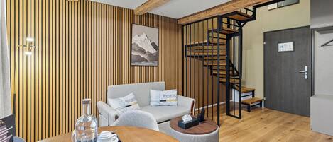 A modern hotel room with a wooden panel wall, a sofa, a round table with a tray of tea and glasses, and a staircase with a wooden railing.