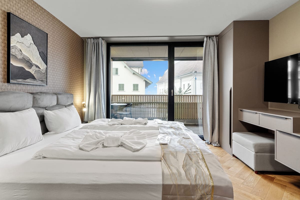 Ett modernt hotellrum med en stor säng, en platt-TV, ett skrivbord och en balkong med utsikt över byggnaderna.