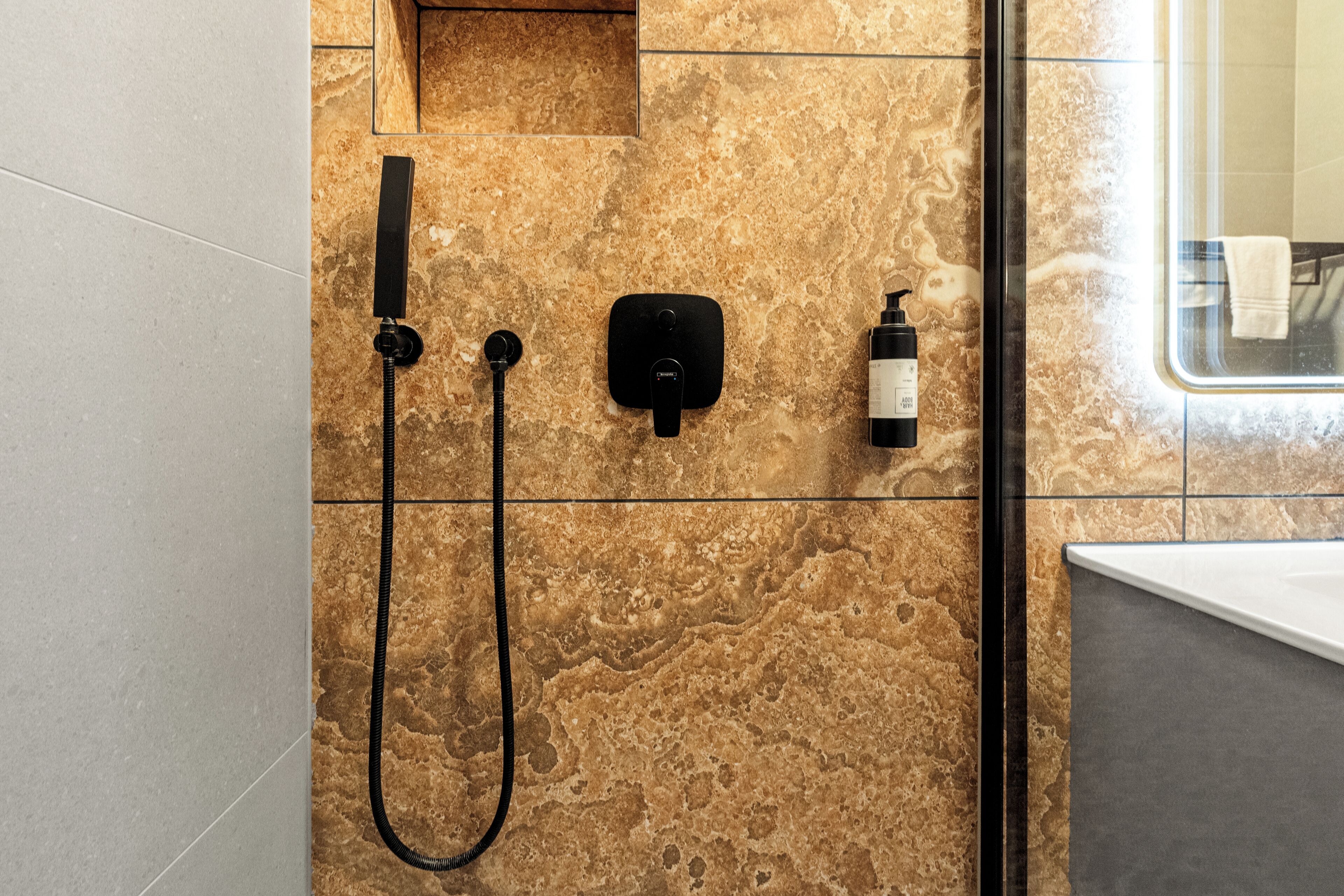 Un baño con ducha, cabezal de ducha manual, panel de control en la pared y una botella de champú.