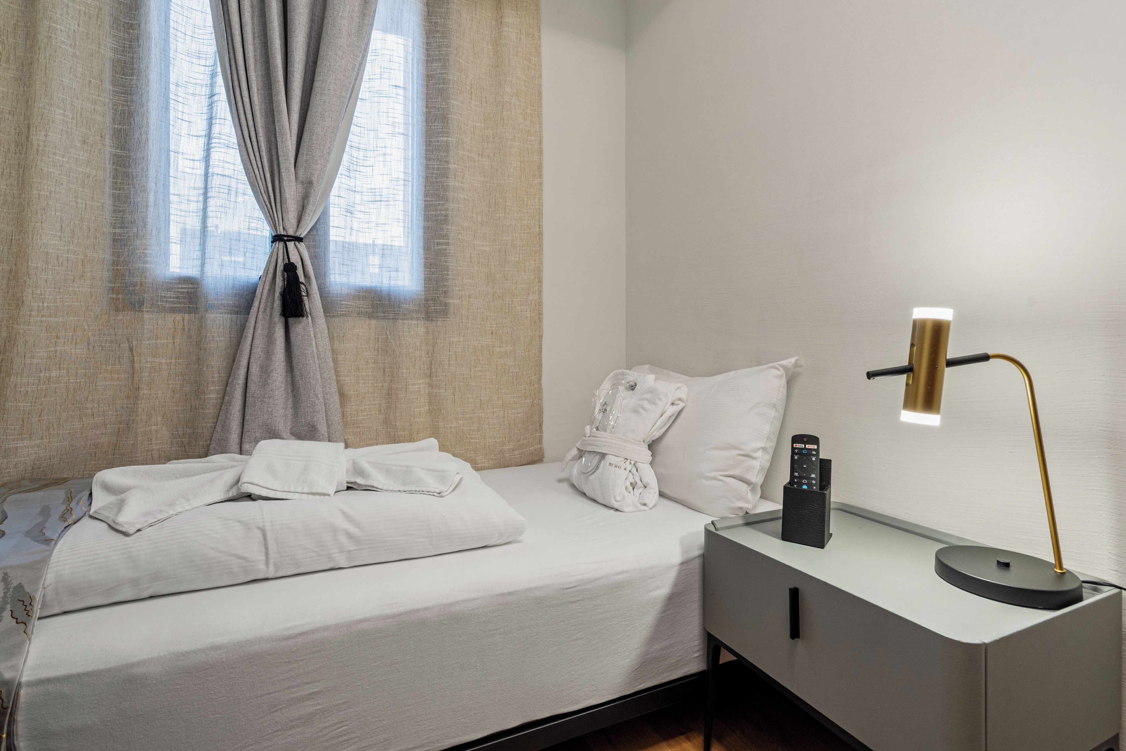Una cama bien tendida con sábanas y toallas blancas, una mesita de noche con lámpara, y una ventana con cortinas de encaje.