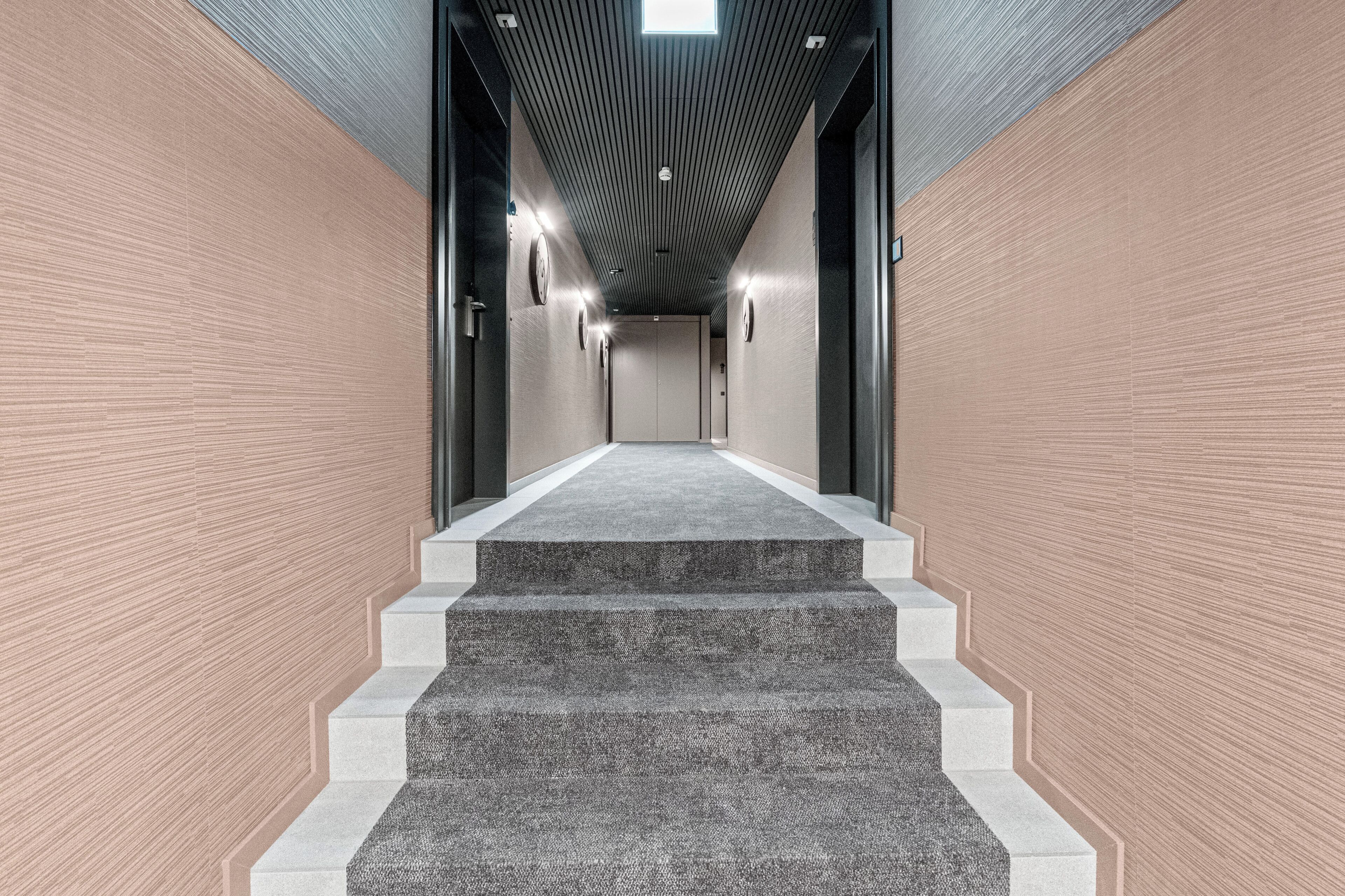 Un pasillo moderno con pared de diseño, una escalera con alfombra e iluminación lineal en el techo.