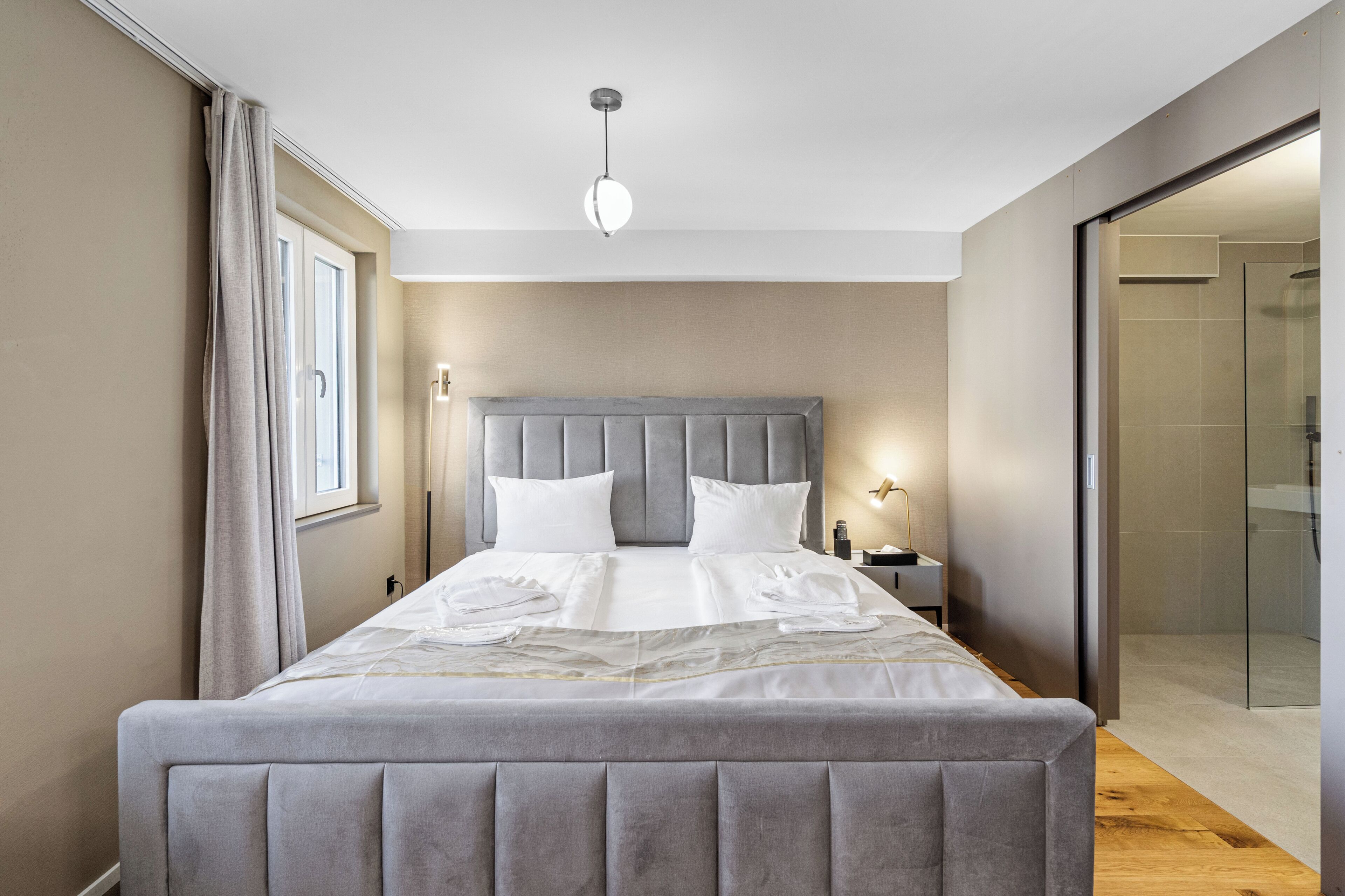 A neatly made bed with a grey headboard and white linens, a bedside table with a lamp, and a glass shower door.