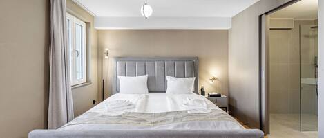 A neatly made bed with a grey headboard and white linens, a bedside table with a lamp, and a glass shower door.