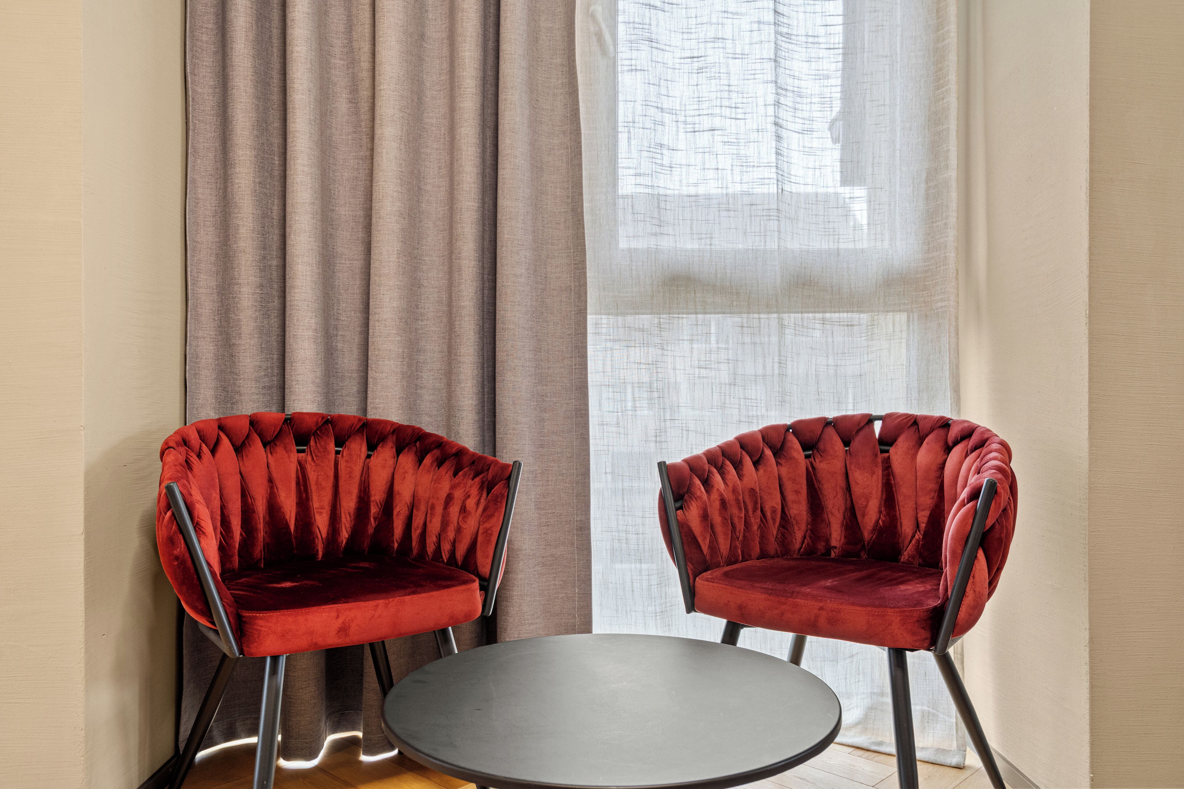 Deux fauteuils en velours rouge, dotés de pieds en métal noir, et une table ronde se trouvent dans une pièce aux murs de couleur beige et à la grande fenêtre ornée de rideaux légers.