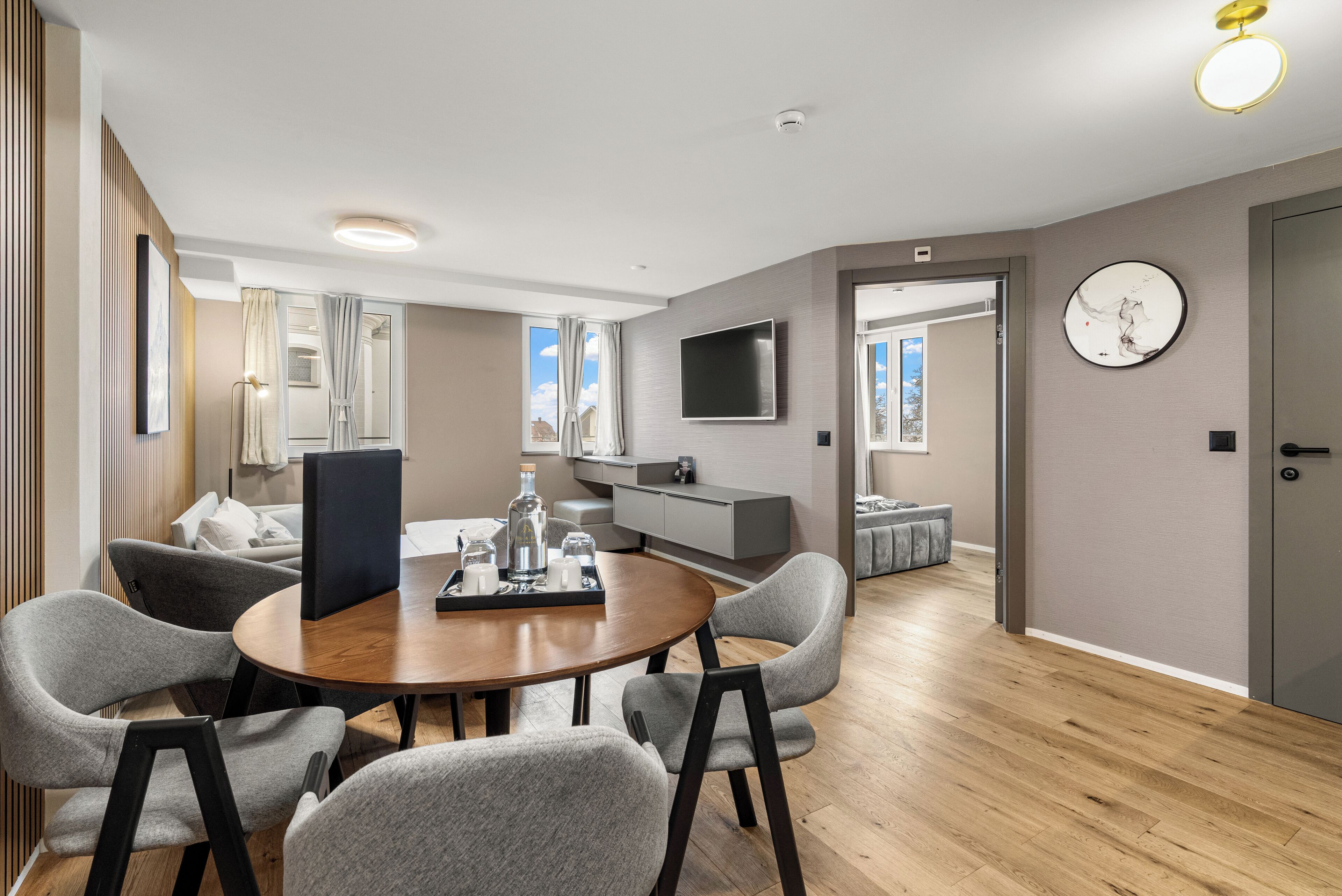 A modern living room with a round wooden table, grey upholstered chairs, a flat-screen TV, and a large wall clock.