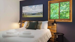 Un dormitorio con cama, almohadas, una mesita de noche, una ventana con vistas a árboles y un cuadro en la pared.