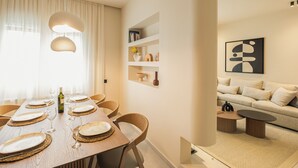 Un coin repas moderne avec une table en bois dressée pour six personnes, entourée de chaises en osier. Un luminaire suspendu est placé au-dessus de la table, et une bouteille de vin y est posée.