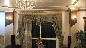 Une pièce avec un lustre, une table ornée d’un vase, deux chaises et une grande fenêtre avec des rideaux.