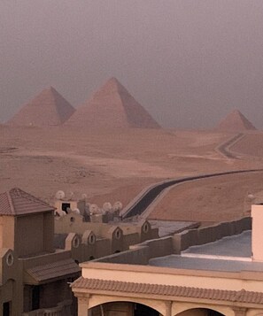 A desert landscape with pyramids in the background and a modern building with a sloped roof in the foreground. - Royal pyramid palace residence (Giza Governorate)