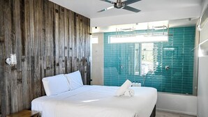 Un dormitorio moderno con cabecera de madera, una cama grande con ropa de cama blanca, un ventilador de techo y vista a una piscina a través de un muro de cristal.
