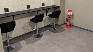 A modern bar area with three black bar stools, a patterned wall, a fire extinguisher, and a ceiling light. - HOTEL GALAXY POD (Koshigaya)