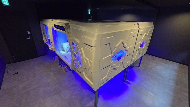 A modern, beige-colored room with a unique geometric design, featuring a bed with a blue-lit headboard and a small window. - HOTEL GALAXY POD (Koshigaya)
