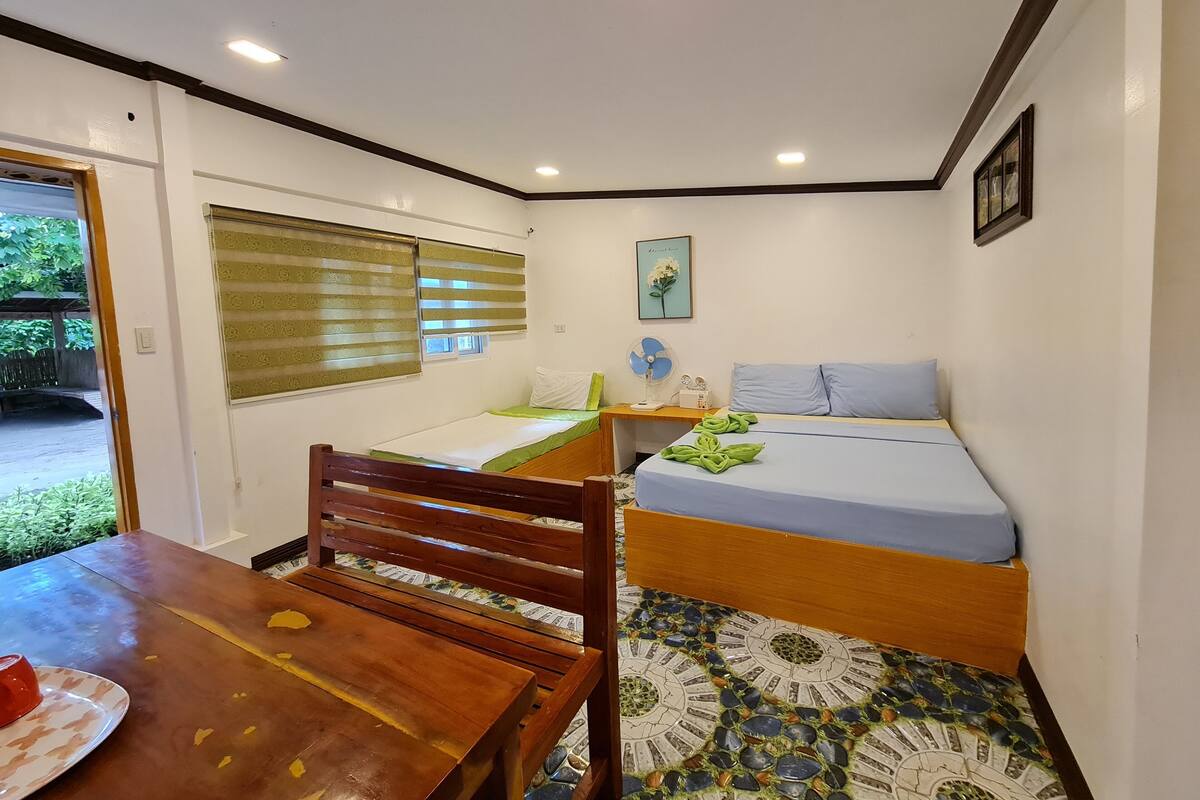 A small, clean room with a bed, a table, and a chair. There is a window with blinds, a picture on the wall, and a fan on a nightstand.