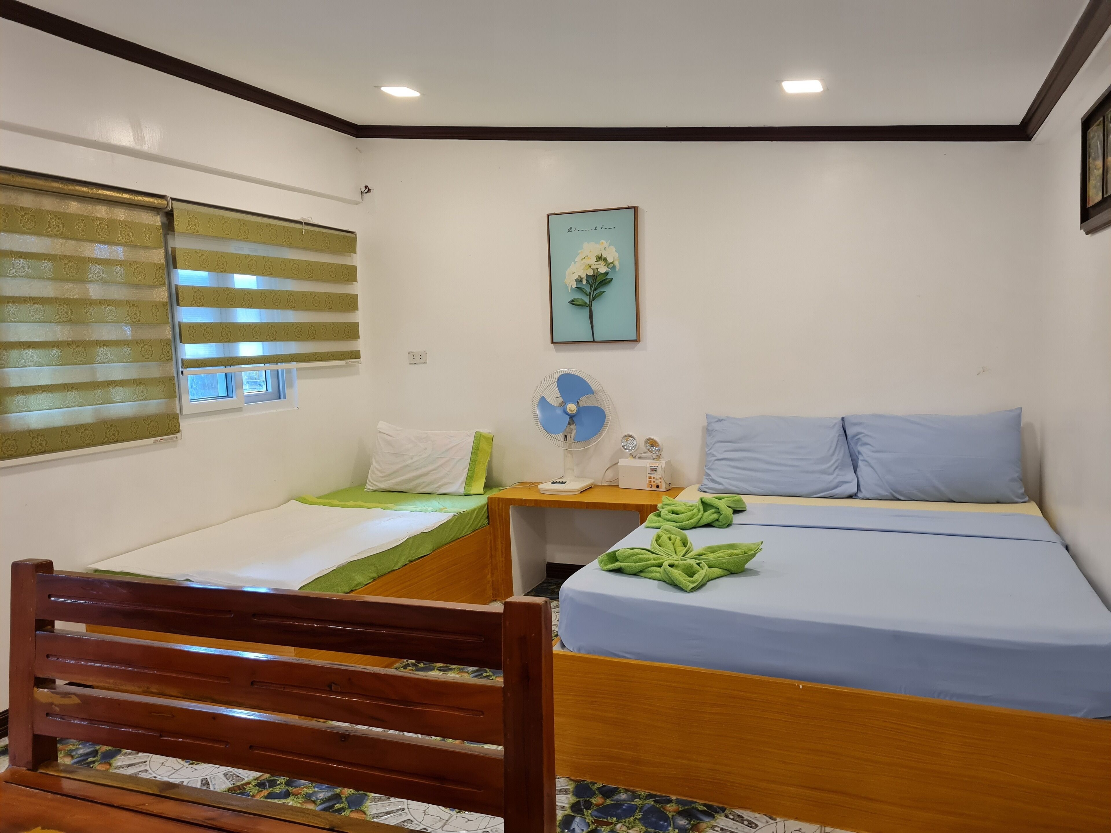 Una habitación pequeña y luminosa con una cama, una silla, una mesita de noche con un ventilador y una ventana con persianas.